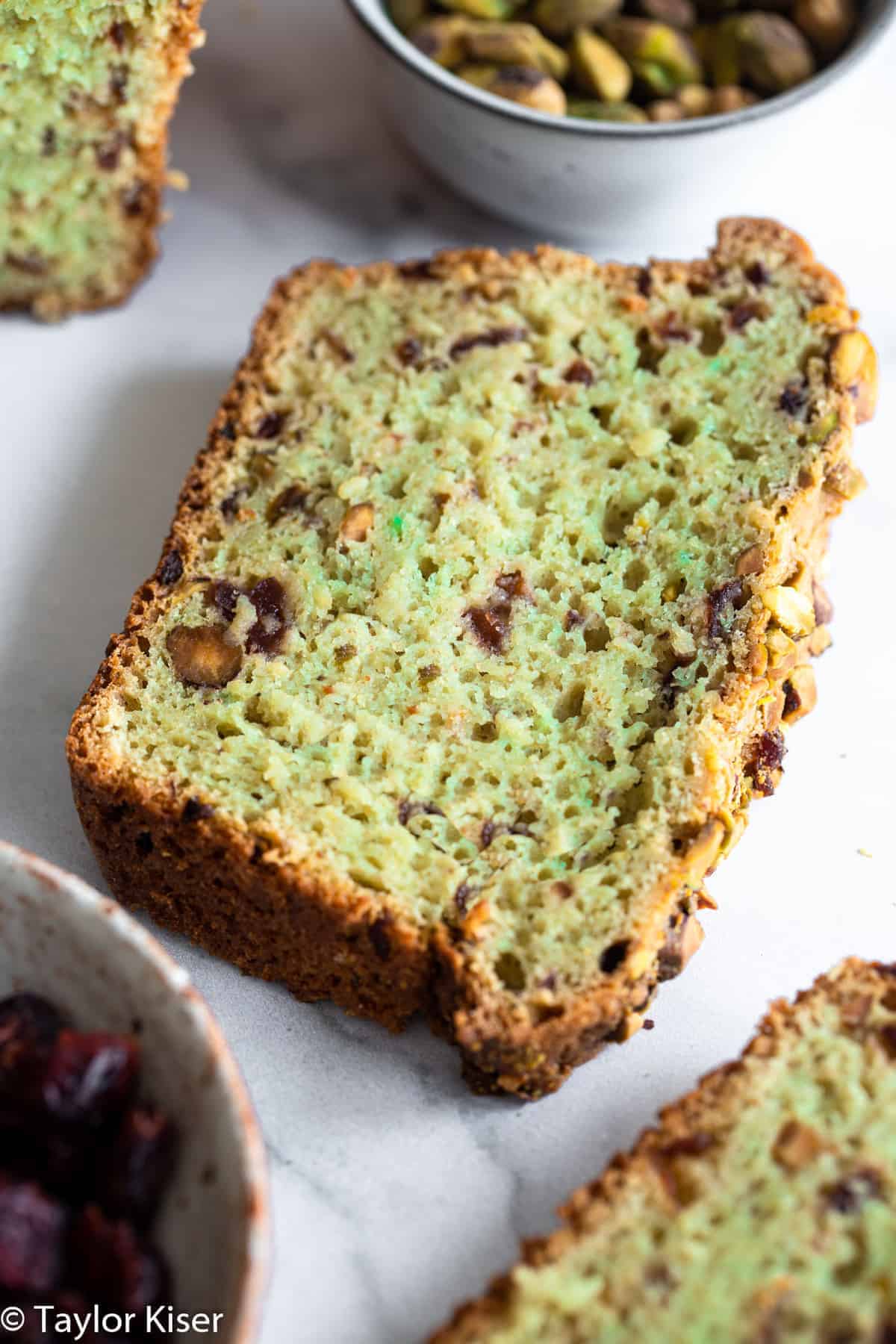 a slice of cranberry bread with a bowl of pistachios beside it